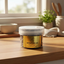 Load image into Gallery viewer, A clear plastic tub of dehydrated emu liver pet treats with a gold label, set against a light-colored wall and surface.