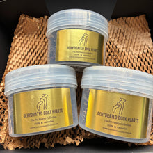 Load image into Gallery viewer, Three clear plastic tubs with gold labels containing dehydrated pet hearts, arranged in a black box lined with brown paper packing material. The labels read 'Dehydrated [animal] Hearts' with a dog silhouette logo and website text.
