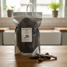 Load image into Gallery viewer, A bag of beef jerky for big dogs, sealed in a silver stand-up pouch with a white label sitting on a wooden bench in the kitchen
